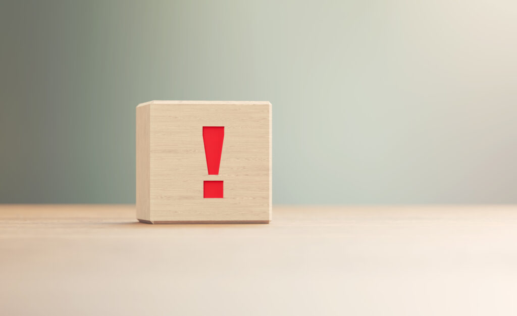 Exclamation point written wood block sitting on wood surface in front of a defocused background. Horizontal composition with copy space.