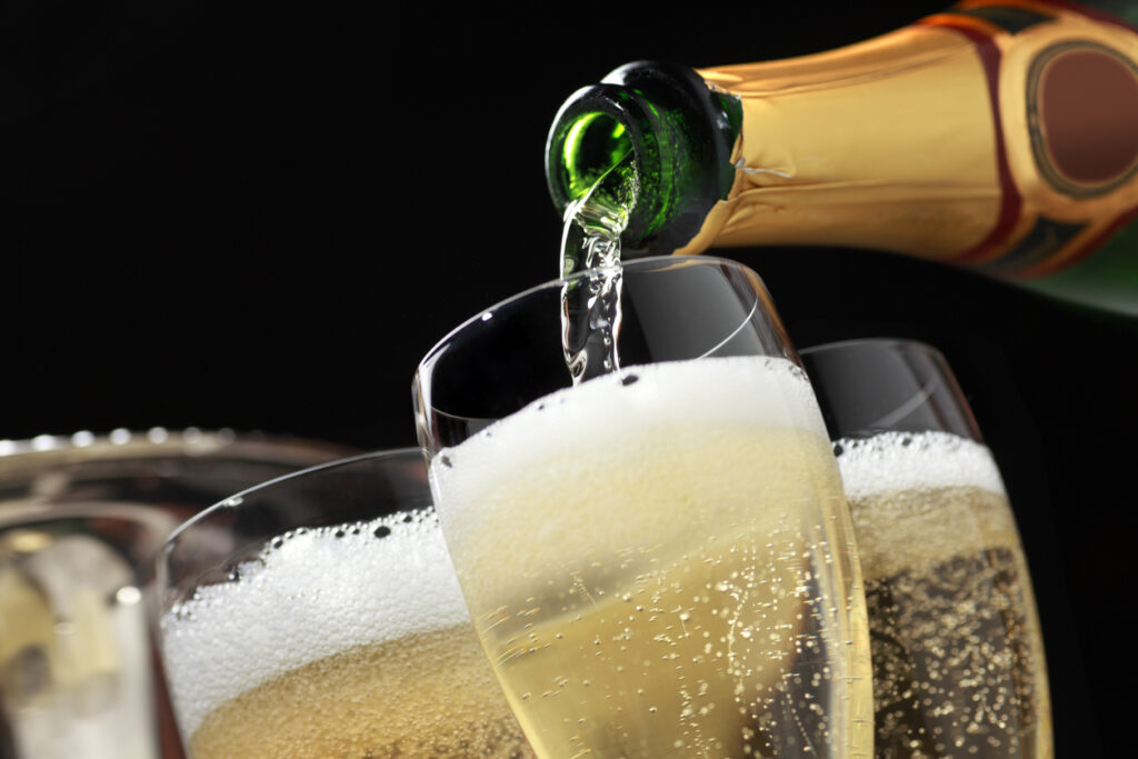 Champagne is being poured into several champagne glasses. The glasses sit in front of the ice bucket that held the champagne bottle as bubbles from the freshly poured champagne can be seen.  The camera is tilted slightly of axis.
