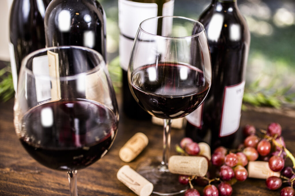 Wine tasting event. Red wine selections on outdoor patio table. Wine bottles in background. Rustic wooden table with grapes. No people.
